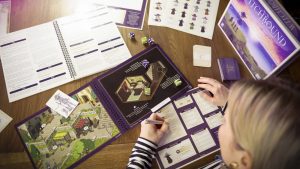 A person looking through the spiral-bound scene book for the solo board game Witchbound, with the game's box sitting nearby on a wooden table.