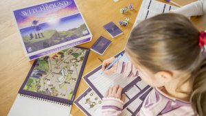 Overhead view of a player reading through the spiral-bound scene book for the Witchbound solo adventure board game, with the game box resting nearby on a wooden table.